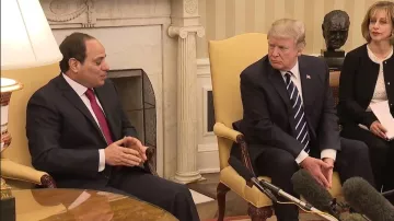 The image shows two men sitting in a formal setting, likely a meeting room in a government building. One man, on the left, is wearing a dark suit and a red tie, appearing to speak or make a point. The other man, on the right, is dressed in a dark suit with a striped tie and is attentively listening or observing. There are microphones positioned between them, indicating that their conversation is being recorded or broadcast. The background features a light-colored wall and a decorative element, possibly a bust or sculpture, along with a warm lamp on the table.