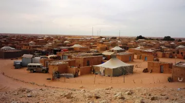 L'image présente un paysage désertique où l'on aperçoit un village de tentes et de maisons en argile. Les structures sont éparpillées dans le sable, entourées d'un terrain aride. Au fond, quelques antennes sont visibles, suggérant une connexion à l'extérieur. Le ciel est nuageux, créant une ambiance calme et légèrement mélancolique.