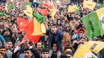 L'image montre une grande foule rassemblée, manifestant avec enthousiasme. Les gens lèvent des drapeaux colorés, dont certains semblent être aux couleurs rouge, jaune et vert. On peut voir un jeune garçon au premier plan exprimant sa joie et son excitation. L'atmosphère est festive, et les participants semblent engagés dans une cause commune, remplis d'énergie et de détermination.