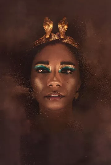 L'immagine presenta un volto femminile con un trucco molto elaborato, caratterizzato da ombretti blu e oro che mettono in risalto gli occhi. La persona indossa una corona decorata con serpenti dorati, che conferisce un aspetto regale e misterioso. Lo sfondo è sfocato e sembra essere avvolto da una polvere dorata, creando un'atmosfera affascinante e drammatica. La luce è calda e accentua la bellezza del viso, rendendo l'immagine suggestiva e intrigante.