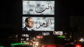 L'image montre un grand panneau d'affichage lumineux à la nuit tombante. Sur ce panneau, on peut voir un portrait d'un homme en costume. Les mots arabes "أنت الأمل" et "أنت" sont écrits en grandes lettres, indiquant un message fort. En bas, il y a une autre image de l'homme. L'environnement est animé, avec des lumières de voitures qui passent en bas, créant un effet de mouvement et une ambiance urbaine nocturne.