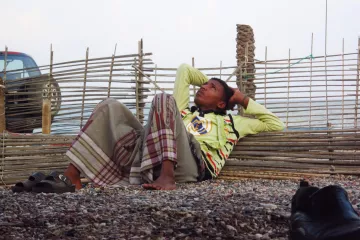 Un garçon allongé, pensif, contre une barrière en bois, sur une plage de gravier.