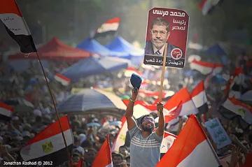 L'image montre une grande foule de manifestants brandissant des drapeaux égyptiens. Au premier plan, un homme lève une pancarte avec le portrait de Mohamed Morsi, qui était le président de l'Égypte. Les participants semblent enthousiasmés et mobilisés, reflétant une atmosphère de rassemblement populaire et d'engagement politique. Le paysage est rempli de tentes et d'une multitude de personnes, créant une impression de solidarité.