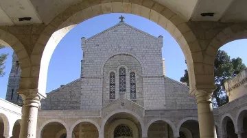 L'image montre un cloître avec des arches stylisées entourant un jardin. Au centre, on peut voir une structure architecturale en pierre, probablement une église ou un monastère, avec un toit en pente et des fenêtres ornées. L'ambiance est paisible, avec une lumière naturelle qui illumine le décor, créant un contraste entre l'ombre des arches et la clarté extérieure. Des éléments de verdure, comme des plantes, ajoutent une touche de vie à l'environnement.