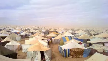 L'image montre un vaste camp de tentes, s'étendant à perte de vue dans un environnement désertique. Les tentes, de diverses tailles et couleurs, sont disposées de manière dense sur le sol sablonneux. Le ciel est nuageux, ce qui donne une atmosphère grise et légèrement menaçante, suggérant peut-être des conditions météorologiques difficiles. On peut également apercevoir de la poussière soulevée par le vent, ajoutant à l'impression de désolation et d'isolement de cet espace.