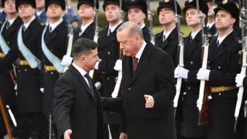 L'image montre deux hommes en discussion. L'un est vêtu d'un manteau noir, tandis que l'autre porte un costume avec une cravate. En arrière-plan, des soldats en uniforme présentent les honneurs, debout et alignés, avec des armes à la main. L'atmosphère semble solennelle, indiquant probablement une rencontre officielle ou un événement diplomatique.