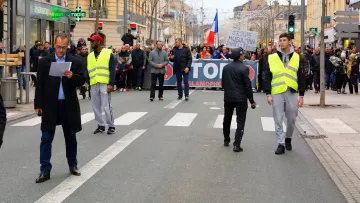 L'image montre une manifestation dans une rue où un groupe de personnes tient une grande banderole avec des slogans. Au premier plan, deux agents de sécurité ou policiers portent des gilets réfléchissants et semblent surveiller la situation. Des manifestants sont visibles à l'arrière, et il y a une ambiance de mobilisation avec des drapeaux. L'environnement urbain se présente avec des bâtiments voisins et une circulation routière réduite à ce moment.