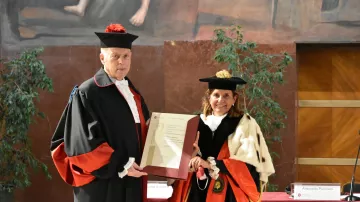L'immagine mostra due persone vestite in abiti accademici formali, tipici per cerimonie di laurea o conferimenti di titoli. Il soggetto a sinistra indossa un mantello nero con dettagli rossi e un cappello, mentre a destra c'è una donna con un abito simile, ma con un mantello e un cappello che contengono diversi elementi decorativi. Entrambi stanno tenendo un documento o un certificato che sembra essere di rilevanza accademica o ufficiale. Sullo sfondo, ci sono piante e un muro decorato, indicando un ambiente formale e celebrativo.