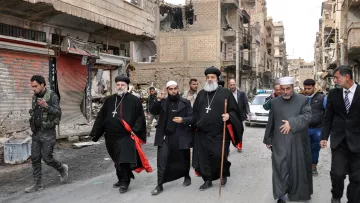 The image shows a group of religious figures walking through a war-torn street. They are dressed in traditional clergy attire, including black robes and crosses, and some are holding staffs. The surroundings are heavily damaged, with buildings showing signs of destruction and debris on the street. In the background, there are a few people and vehicles, indicating a somber atmosphere in a once-bustling area. The overall scene reflects the impact of conflict on the community and highlights the presence of religious leadership amidst the destruction.