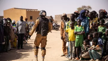 L'image montre un soldat en uniforme militaire, portant un casque et un sac à dos, se tenant face à une foule. Les personnes dans la foule sont de différentes tranches d'âge, allant des enfants aux adultes, et semblent observer le soldat avec attention. Le décor évoque une ambiance de village rural, avec des bâtiments simples et un sol en terre battue. Les couleurs de l'uniforme du soldat contrastent avec celles des vêtements des membres de la foule. L'atmosphère générale semble être celle d'une interaction, peut-être dans le cadre d'une mission de sécurité ou d'assistance.