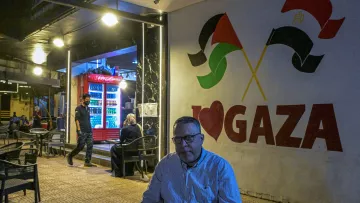 Un homme assis, devant un mural "I ❤️ GAZA", avec des drapeaux palestiniens en arrière-plan.