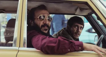 Deux hommes dans une voiture, l'un avec des lunettes de soleil, l'autre semble inquiet.