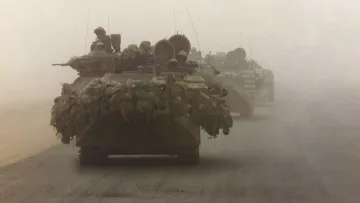 L'image montre des véhicules militaires, probablement des chars, se déplaçant dans un environnement poussiéreux et brumeux. On peut voir des soldats à bord des chars, et ces derniers semblent être chargés avec des équipements. L'atmosphère est chargée, créant une impression de tension et d'intensité, probablement en lien avec une situation de conflit ou d'opération militaire.