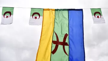 L'image montre plusieurs drapeaux suspendus. Au centre, il y a un grand drapeau de couleur verte avec un symbole rouge au centre, probablement représentant le mouvement amazigh. De chaque côté, il y a de plus petits drapeaux, qui semblent être le drapeau algérien, avec les couleurs verte et blanche et le croissant et l'étoile rouges. L'ensemble est accroché sous un ciel nuageux, créant une ambiance festive ou symbolique.