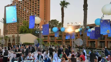 The image depicts an outdoor event, likely a formal gathering or dinner party set up in a picturesque location. Tables are elegantly arranged with white tablecloths and chairs, and guests are dressed in formal attire, engaging in conversation. The ambiance is enhanced by decorative blue flags, likely representing the European Union, and hanging lanterns that illuminate the scene. Palm trees are visible in the background, adding a tropical feel, while a hotel or building looms in the background. The atmosphere appears lively and festive, suggesting a celebration or special occasion.