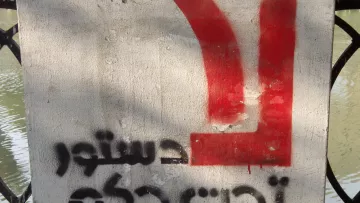 L'image montre un panneau sur un mur en béton, avec un fond gris. Sur la partie supérieure, on peut voir un symbole ou une lettre en rouge. En dessous, des mots sont écrits en arabe, et le texte semble faire référence à un sujet politique, évoquant un appel ou une déclaration. Le tout est en contraste avec l'environnement aquatique en arrière-plan.