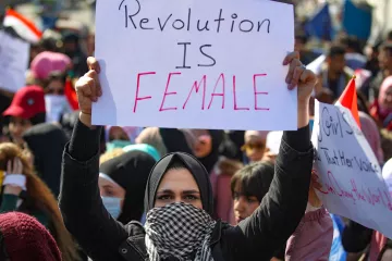 L'immagine mostra una manifestazione con molte persone riunite. In primo piano, una donna indossa un hijab e tiene in alto un cartello su cui è scritto "Revolution is Female". Sullo sfondo si vedono altre persone, alcune delle quali portano bandiere e cartelli, creando un'atmosfera di mobilitazione e protesta. La scena è caratterizzata da un forte senso di unità e determinazione.