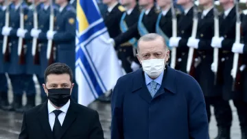 The image shows two men standing side by side in a formal setting, likely during a military ceremony or event. One man is wearing a black face mask and dark suit, and the other is in a blue coat and also wearing a mask. In the background, there is a formation of soldiers in uniform holding rifles, and a flag is prominently displayed, suggesting a significant occasion. The overall atmosphere appears solemn and official.