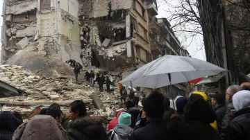 L'immagine mostra una scena dopo un terremoto devastante. Si può vedere un edificio gravemente danneggiato, con pareti crollate e macerie sparse. Alcuni soccorritori stanno cercando di recuperare persone intrappolate tra i detriti. Una folla di persone osserva da una certa distanza, alcune delle quali sono riparate sotto ombrelli, probabilmente a causa della pioggia. L'atmosfera è caratterizzata da tensione e preoccupazione, mentre le squadre di soccorso lavorano per salvare vite.