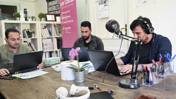 L'image montre trois hommes assis autour d'une table en bois, chacun travaillant sur un ordinateur portable. Ils semblent concentrés sur leurs tâches. Sur la table, on peut voir des objets tels qu'un casque, des stylos et une plante en pot. À l'arrière-plan, il y a des affiches et des livres, ce qui donne une ambiance de bureau ou d'espace de travail collaboratif. Un microphone est également visible sur la table, ce qui pourrait indiquer qu'ils sont en train d'enregistrer ou de diffuser quelque chose.