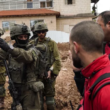 L'image montre des soldats en uniforme militaire, armés, entourés par un groupe de personnes. Certains sont en tenue d'urgence, tandis que d'autres semblent discuter avec les soldats. En arrière-plan, on voit des bâtiments et un terrain boueux. L'ambiance est tendue.