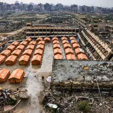 L'image montre un paysage de destruction, avec des bâtiments en ruines en arrière-plan. Au premier plan, des tentes orange sont disposées en rangées sur un terrain dévasté, témoignant d'une situation de crise ou de catastrophe.