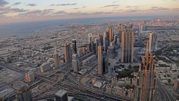 L'image montre un panorama impressionnant d'une ville moderne, probablement Dubai. On y voit de nombreux gratte-ciels élégants s'élevant vers le ciel, ainsi qu'un réseau complexe de routes et d'autoroutes serpentant à travers la métropole. À l'arrière-plan, on aperçoit la mer et des paysages urbains qui s'étendent à perte de vue. Le ciel présente des teintes douces de bleu et d'orange, témoignant d'un coucher de soleil, ce qui ajoute une ambiance tranquille à la scène urbaine animée.