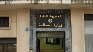 L'image montre un bâtiment avec une façade en pierre, portant une inscription en arabe et en français. On y voit un soldat en uniforme, armé, qui se tient devant l'entrée du bâtiment. Le soldat semble en position de garde, et à côté de lui, on peut remarquer des éléments architecturaux typiques des bâtiments gouvernementaux. L'inscription indique que l'édifice est le ministère des Finances du Liban.