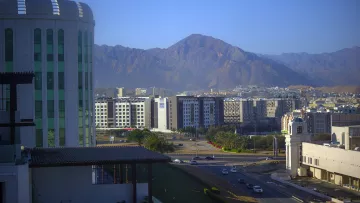 La imagen muestra un paisaje urbano con una mezcla de edificios modernos y montañas al fondo. A la izquierda, se puede ver un edificio de gran altura con un diseño contemporáneo. En el centro, hay una serie de edificios de varios pisos, que parecen ser parte de un área urbana. Al fondo, se aprecian montañas con una forma escarpada y un color terroso, indicando un ambiente montañoso. La luz del sol ilumina la escena, generando un ambiente claro y fresco. En primer plano, hay una carretera que conecta los diferentes sectores de la ciudad.