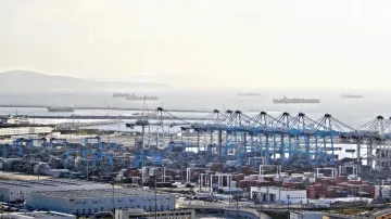 L'image montre un port maritime avec des conteneurs empilés, des grues de chargement et plusieurs navires au large. On peut voir des bâtiments et des installations portuaires. L'horizon est légèrement brumeux, créant une atmosphère calme, et des bateaux de différentes tailles sont visibles sur la mer. L'ensemble dépeint une scène de transport maritime et de logistique.