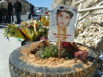 L'image montre un mémorial pour un enfant, avec une photo de lui affichée sur un panneau en bois planté dans un pneu. Autour du panneau, il y a des fleurs multicolores et de la terre. En arrière-plan, on peut voir des personnes rassemblées et un paysage de pierre. Cela semble être un hommage émouvant à un enfant décédé.