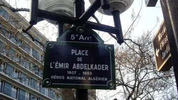 L'image montre un lampadaire avec plusieurs globes lumineux, sur lequel est fixée une plaque signalétique. Cette plaque indique "Place de l'Émir Abdelkader", accompagnée des dates 1807-1883 et de la mention "Héros National Algérien". Le tout se situe dans le cinquième arrondissement, comme le précise le petit panneau en haut. En arrière-plan, on peut apercevoir des bâtiments typiques de la ville.