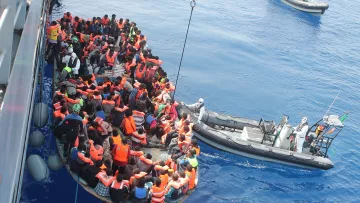 L'image montre un groupe de personnes dans une embarcation sur une mer calme. La plupart des individus portent des gilets de sauvetage orange, indiquant qu'ils sont en situation de danger. À proximité, une autre petite embarcation semble aider le groupe, probablement une équipe de secours. L'atmosphère générale évoque des enjeux humanitaires, liés à la migration et aux sauvetages en mer. Le ciel est clair et l'eau est d'un bleu profond.