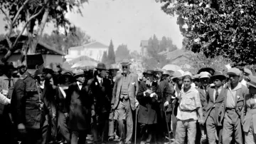 L'image montre un groupe de personnes rassemblées en extérieur, avec une scène animée. Au centre, une figure éminente se déplace, soutenue par une canne. De nombreuses personnes autour d'elle portent des chapeaux et tiennent des parasols, probablement pour se protéger du soleil. Des hommes en costume se distinguent parmi la foule, et l'environnement semble être urbain, avec des bâtiments en arrière-plan. La scène dégage une atmosphère d'importance ou de célébration.