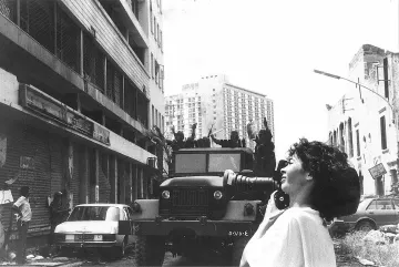 L'image montre une scène urbaine où une femme tient un appareil photo, prenant probablement des clichés d'une situation troublée. En arrière-plan, on peut voir un véhicule militaire sur une route, avec plusieurs personnes en uniforme visible sur le camion. Les bâtiments autour semblent en mauvais état, avec des stores métalliques fermés. L'atmosphère dégage une impression de tension ou de conflit. La femme, avec des cheveux bouclés, semble concentrée sur sa prise de vue, capturant un moment significatif dans un contexte difficile.