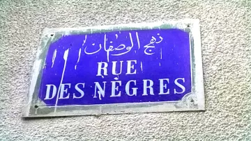 تصویر یک تابلو خیابان را نمایش می‌دهد که دارای نوشته‌هایی به زبان‌های عربی و فرانسوی است. زمینه تابلو به رنگ آبی است و نوشته‌ها به رنگ سفید. در قسمت بالای تابلو، متن عربی وجود دارد که در پایین آن، نام خیابان به زبان فرانسوی (Rue des Nègres) نوشته شده است. تابلو به نظر می‌رسد که قدیمی است و ممکن است در معرض عوامل جوی قرار داشته باشد، زیرا آثاری از کثیفی یا ساییدگی بر روی آن مشاهده می‌شود.
