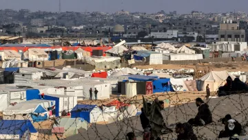 L'image montre un camp de réfugiés avec de nombreuses tentes de différentes couleurs, principalement blanches et bleues, s'étendant sur un terrain désertique. À l'arrière-plan, on aperçoit une ville avec des bâtiments. On peut également distinguer des personnes assises, ainsi que des barbelés entourant le camp, ce qui souligne une ambiance de sécurité précaire et d'incertitude. L'ensemble évoque une situation difficile et des conditions de vie sommaires.