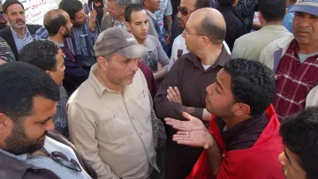 L'image montre un groupe de personnes rassemblées dans un endroit public, probablement lors d'une manifestation ou d'un rassemblement. Plusieurs hommes se tiennent ou se parlent les uns aux autres, affichant des expressions variées qui semblent indiquer une discussion animée. Certains portent des vêtements décontractés et il y a une ambiance de dialogue, avec des gestuelles qui suggèrent l'échange d'opinions. En arrière-plan, on peut apercevoir d'autres personnes qui regardent la scène. L'atmosphère semble engageante et dynamique.