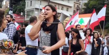 Foule en manifestation, femme au mégaphone, drapeaux libanais en arrière-plan.