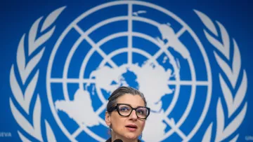 L'image montre une femme parlant devant un microphone, avec un fond représentant le logo des Nations Unies. Elle porte des lunettes et a les cheveux attachés. Son expression semble concentrée, et elle est probablement en train de faire une déclaration ou de répondre à des questions. Le décor bleu est typique des conférences de presse internationales.