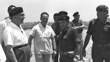 The image appears to show a group of men in a casual outdoor setting, likely involved in a military or political context. Some individuals are dressed in military uniforms, while others wear casual or formal attire. The atmosphere seems informal as they engage in conversation, suggesting a moment of collaboration or discussion. The background features a rural landscape, possibly indicating a visit to a site of significance.