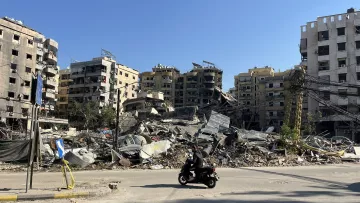 L'image montre une scène de destruction urbaine, où plusieurs bâtiments sont partiellement effondrés et en ruines. On peut voir des décombres éparpillés au sol, tandis qu'une personne à moto passe devant. Le ciel est dégagé, laissant transparaître un jour ensoleillé, contrastant avec le désastre visible. La scène évoque un fort sentiment de désolation et de dégâts, typique de zones touchées par des conflits ou des catastrophes.