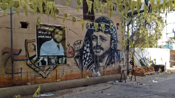 L'image montre un mur décoré d'une grande fresque représentant un homme avec un keffieh, typique de la culture arabe. À côté, il y a des affiches avec des portraits et des slogans. Des drapeaux jaunes pendent également, créant une ambiance festive ou militante. Le sol semble être en désordre avec divers objets autour, indiquant peut-être un événement ou une célébration récente. Les couleurs vives des décorations contrastent avec le mur beige.