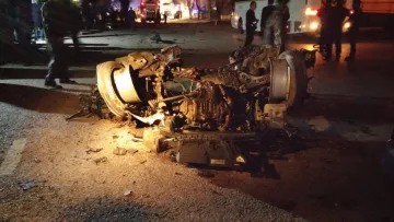 L'image montre un véhicule gravement endommagé, probablement retourné sur le côté. On peut voir des débris éparpillés sur la route, indiquant un accident violent. La scène est éclairée par des lumières, suggérant la présence de services d'urgence ou de témoins. Le contexte semble être nocturne, ce qui accentue les dégâts visibles sur le véhicule.