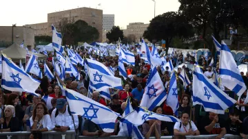 L'image montre une grande foule de personnes rassemblées, brandissant des drapeaux israéliens. Les manifestants semblent réunis pour une occasion particulière, avec une ambiance dynamique. En arrière-plan, on peut apercevoir des bâtiments et des structures urbaines, tandis que des tentes sont installées, peut-être pour fournir des services aux participants. L'ensemble illustre une manifestation significative, avec une forte présence de la couleur bleue et blanche des drapeaux.