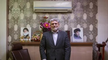 The image shows a man standing in an office setting. He is dressed in a formal suit and has a friendly expression. Behind him, there are two portraits displayed on the wall, likely of influential figures. The office has decorative elements, including a floral arrangement and a neatly organized desk with various items. The overall atmosphere conveys professionalism and authority.