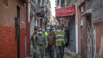 الصورة تظهر زقاقًا ضيقًا في إحدى المدن، حيث يسير مجموعة من الأشخاص. يرتدي بعضهم زيًا رسميًا، مما يشير إلى أنهم من عناصر الأمن أو الشرطة، ويبدو أنهم يقومون بدورية أو لجنة تفتيش. تظهر خلفهم مبانٍ قديمة وممرات ضيقة، مع بعض العلامات التجارية والستائر الملونة. الأجواء توحي بنشاط وحركة في المكان.