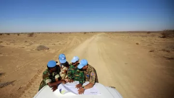 L'image présente un groupe de personnes, probablement des soldats de la paix, qui se penchent sur une carte étalée sur le toit d'un véhicule. Ils portent des casquettes de l'ONU et des uniformes militaires. L'arrière-plan montre un paysage désertique, avec un sol sablonneux et peu de végétation, sous un ciel ensoleillé et dégagé. La scène semble se dérouler dans une région aride, éloignée, où ces individus discutent des informations cartographiques.