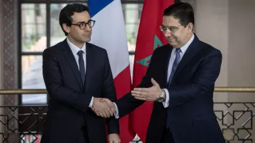 L'image montre deux hommes en costume, se serrant la main dans un cadre officiel. À l'arrière-plan, on peut voir un drapeau français à gauche et un drapeau marocain à droite. L'atmosphère semble être celle d'une rencontre diplomatique ou d'une conférence de presse, soulignant une interaction entre les deux pays.