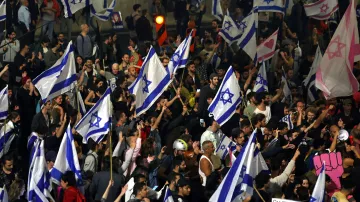 L'image montre une grande foule rassemblée, où des personnes tiennent des drapeaux israéliens. L'atmosphère est festive et engagée, avec des signes et des expressions de mobilisation. On peut observer une diversité de participants, certains portant des cris ou des slogans. La scène semble se dérouler lors d'une manifestation ou d'un rassemblement public.